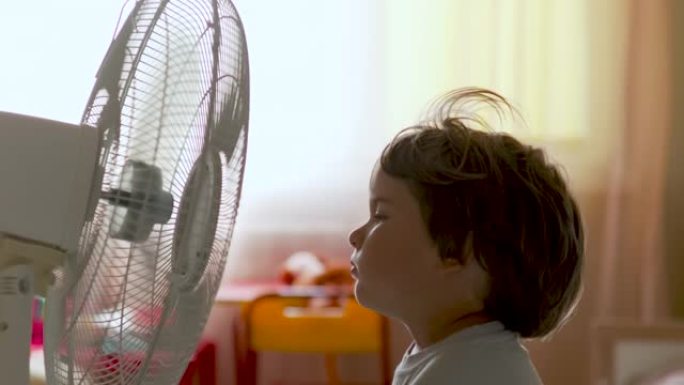 男孩站在范面前。暑假在家享受电风扇带来的凉风的孩子。用电风扇冷却器冷却自己的通风机前受热高温。