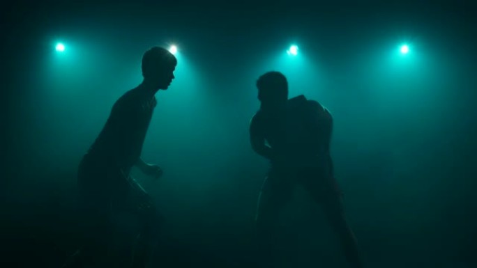 两个家伙的轮廓篮球运动员练习进攻，运球和防守技巧。高大的竞争对手在黑暗的工作室里用烟雾和蓝光竞争篮球