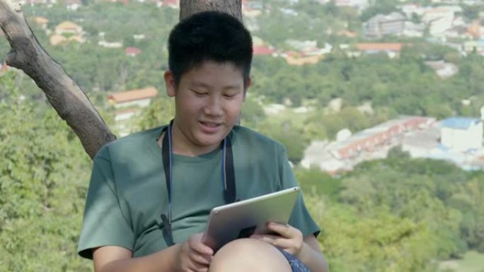 快乐的亚洲男孩使用数字平板电脑在树下的山顶，生活方式的概念。