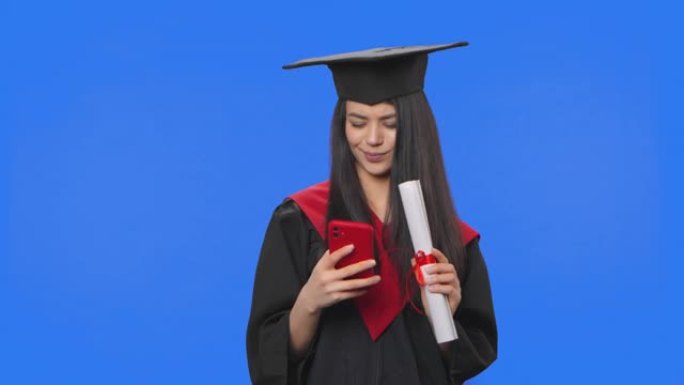 毕业服装女学生肖像，拿着文凭，用手机自拍。年轻女子在工作室摆姿势，蓝屏背景。特写。慢动作准备59.9