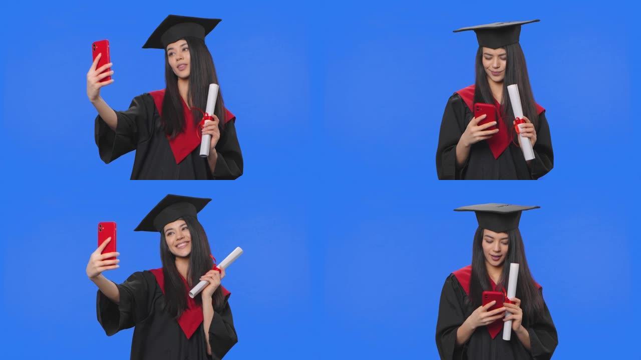 毕业服装女学生肖像，拿着文凭，用手机自拍。年轻女子在工作室摆姿势，蓝屏背景。特写。慢动作准备59.9
