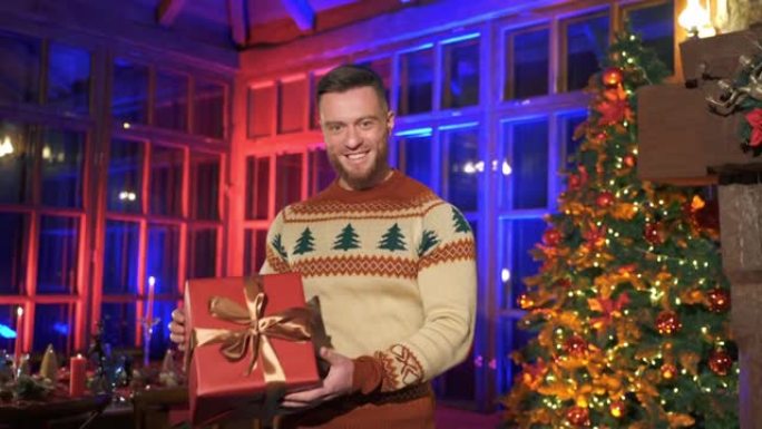 微笑的人提供新年礼物。一个穿着针织圣诞毛衣的英俊男子的肖像，在圣诞装饰的房间背景上拿着一个带礼物的盒