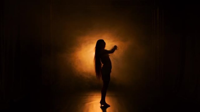 性感的年轻女子在明亮的黄光背景下，在黑暗的烟熏工作室里跳舞和挥动长发。一位穿着短裙的女性正以慢动作摆