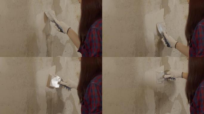 房间里戴着手套在墙上涂油灰的年轻女子的特写镜头。女性用油灰刀抹灰墙，准备在公寓里绘画。维修工作的概念