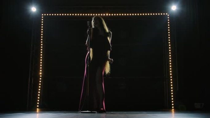 一位穿着红色长裙的别致女独奏者的仰视图，在空荡荡的舞台上唱着复古的麦克风。长头发的Artisca在黑