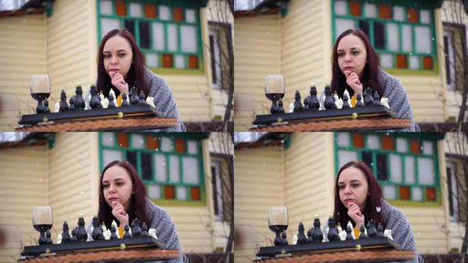 年轻女子下棋，在院子里喝红酒。冬季，女性穿着灰色格子裹着酒精，坐在街上玩棋盘游戏