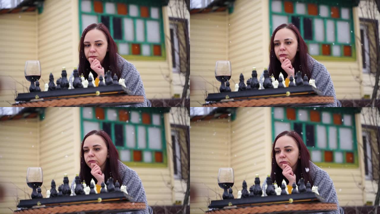 年轻女子下棋，在院子里喝红酒。冬季，女性穿着灰色格子裹着酒精，坐在街上玩棋盘游戏