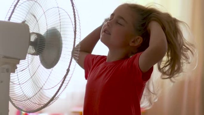 站在范面前的女孩。暑假在家享受电风扇带来的凉风的孩子。用电风扇冷却器冷却自己的通风机前受热高温。
