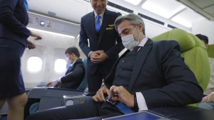 低角度视角: 高加索高级商人戴着防护面罩系好安全带，作为以下航空管家检查乘客的建议，登机准备起飞，踏