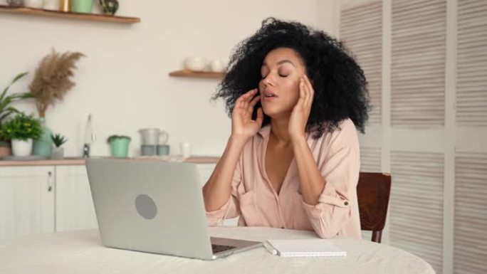 可爱的非洲裔美国女人专注于在笔记本电脑上思考和打字，累了擦太阳穴，不知道。黑人女性坐在明亮厨房的桌子