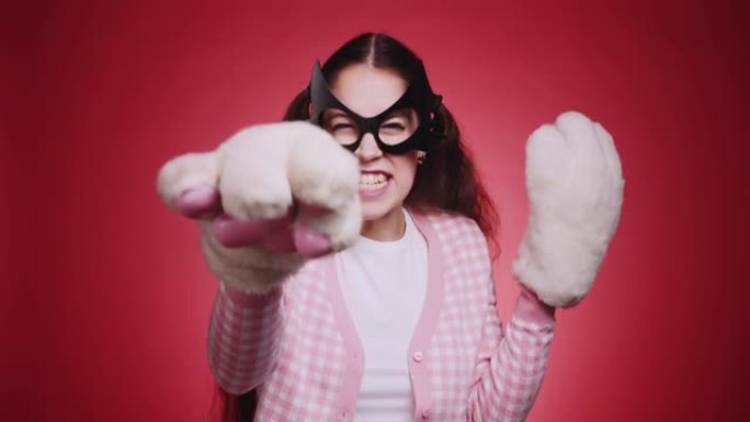 可爱的女孩，戴着猫面具和粉红色的衣服，毛绒蓬松的爪子在红色背景的镜头前调情猫的手势。猫女搞笑角色扮演