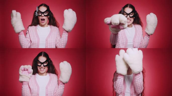 可爱的女孩，戴着猫面具和粉红色的衣服，毛绒蓬松的爪子在红色背景的镜头前调情猫的手势。猫女搞笑角色扮演