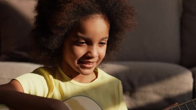 肖像小非洲裔美国女孩在明亮的阳光下弹吉他。十几岁的女孩坐在大厅的地板上，背景是明亮美丽的房间内部。慢