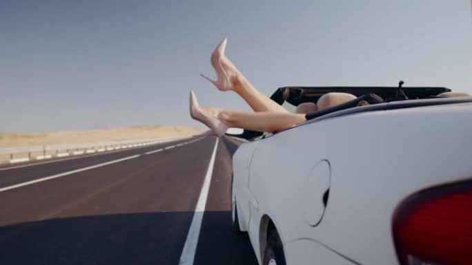女人躺在后座的敞篷车上，在空荡荡的道路背景下，摆动的双腿特写镜头。在空荡荡的高速公路上，用白色敞篷车