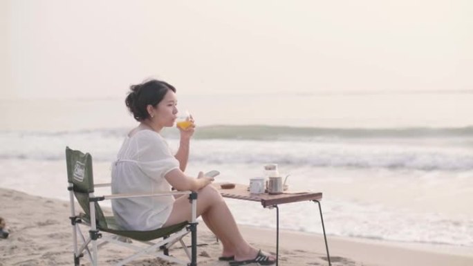 年轻女子在海滩上放松和使用智能手机时喝橙汁。