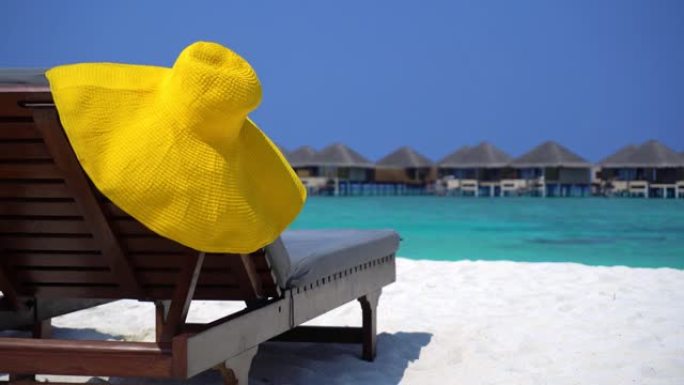 热带沙滩上日光浴浴床上的遮阳帽，以水上别墅为背景。旅游目的地