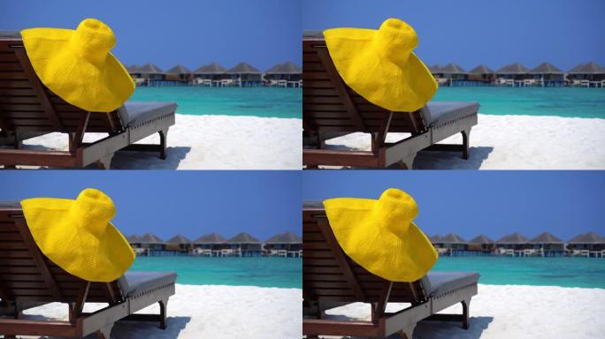 热带沙滩上日光浴浴床上的遮阳帽，以水上别墅为背景。旅游目的地