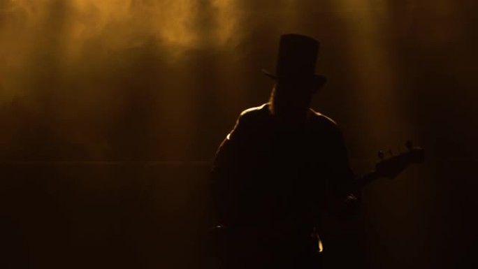 摇滚音乐家在烟雾和明亮的黄色霓虹灯中弹奏低音吉他的动态表演。穿着皮大衣和帽子的人表演现场音乐会。剪影