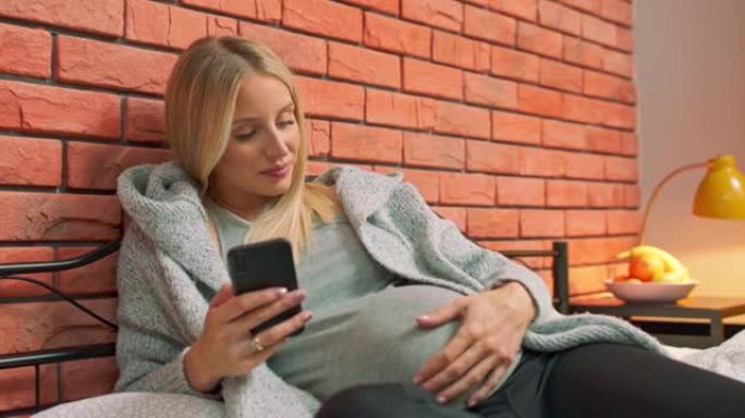 快乐的孕妇年轻女子通过使用智能手机聊天并发短信给丈夫或家人，她的婴儿或胎儿身体健康，身体强壮，几个月