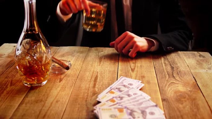 穿着黑色西装的商人正在用威士忌在玻璃旁边的木桌上扔一捆钱