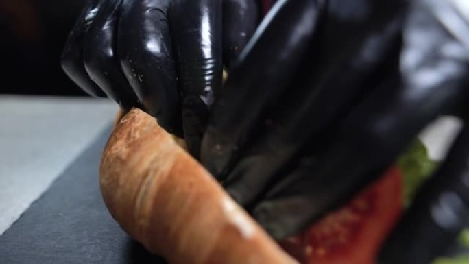厨师的手戴着黑手套准备羊角面包三明治的特写镜头。快速早餐。将切达干酪放在培根散装的番茄上。