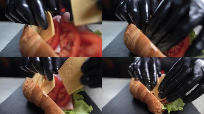 厨师的手戴着黑手套准备羊角面包三明治的特写镜头。快速早餐。将切达干酪放在培根散装的番茄上。