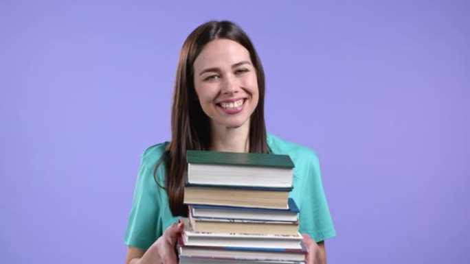 学生在工作室的紫罗兰色背景下从图书馆拿着一堆大学书籍。女人微笑着，她很高兴毕业。