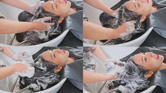 亚洲年轻女子躺在沙龙洗发床上，由发型师在发廊洗头。美发师按摩和用水和洗发水洗头时，美丽的顾客交谈和放