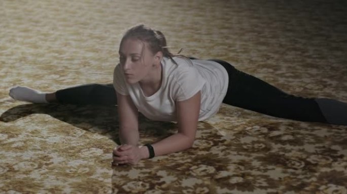 体操女孩坐在体操垫上的麻线上倾斜。在一个明亮的聚光灯下，在黑暗的健身房里穿着运动服伸展体操运动员。晚