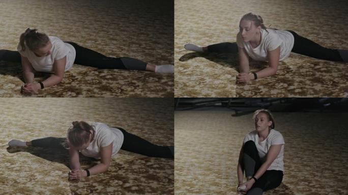 体操女孩坐在体操垫上的麻线上倾斜。在一个明亮的聚光灯下，在黑暗的健身房里穿着运动服伸展体操运动员。晚