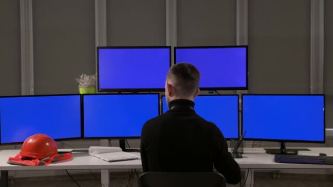 一位工程师坐在许多电脑屏幕前。桌子上是橙色的头盔和背心。从后面看。他浏览记录和电话。