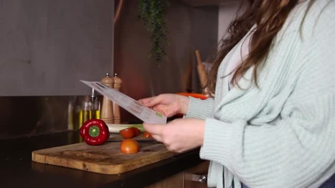 快乐的年轻女子正在现代家庭厨房里一步一步用纸制作素食食谱。微笑的女性正在烹饪健康的素食沙拉，使用食谱
