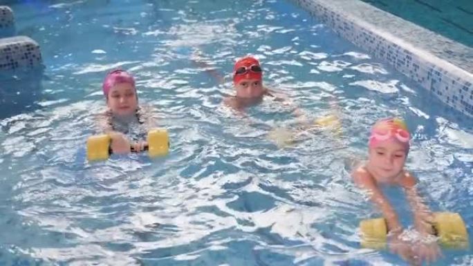 游泳课中的三个小孩使用泡沫哑铃在游泳池游泳