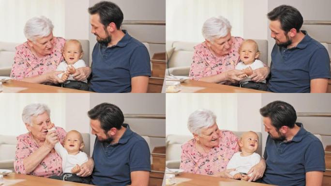 老年妇女将孙子抱在腿上，她的儿子坐在她旁边，他们在餐桌旁度过时光，妇女正在和孩子玩耍，她举起手，孩子