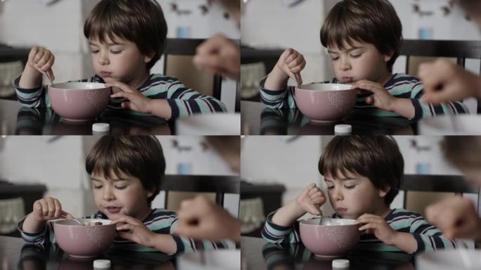 孩子吃玉米片和牛奶。饥饿的小男孩坐在厨房的餐桌上，从盘子里吃早餐玉米麦片。健康营养薄片。早餐吃早餐的