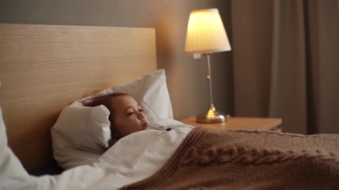 可爱的生病的小女孩躺在床上在家用体温计测量温度。