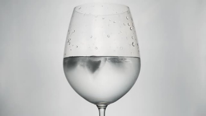 特写清澈的水在生动的玻璃葡萄酒与白色背景和照明在工作室与冰块旋转运动显示清凉的水饮料和透明溢出的冷冻