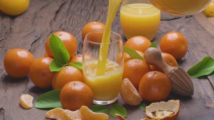 将新鲜的橘子从水器倒入玻璃杯中。周围是成熟的橘子，叶子在旧的木制表面上。