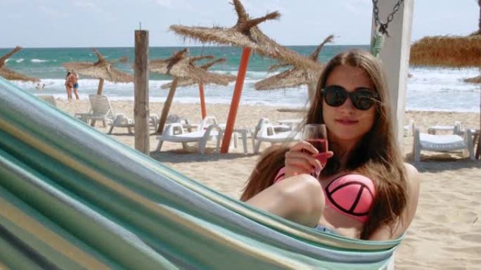 一个年轻女子坐在吊床上，拿着一杯玫瑰酒，在海滩上享受夏日的阳光。暑假假期旅行概念。4k慢动作