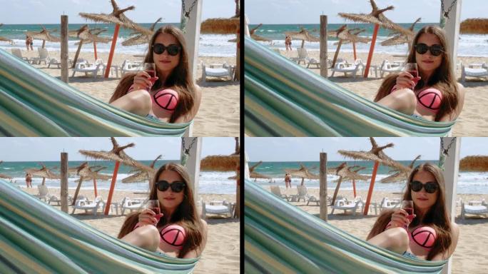 一个年轻女子坐在吊床上，拿着一杯玫瑰酒，在海滩上享受夏日的阳光。暑假假期旅行概念。4k慢动作