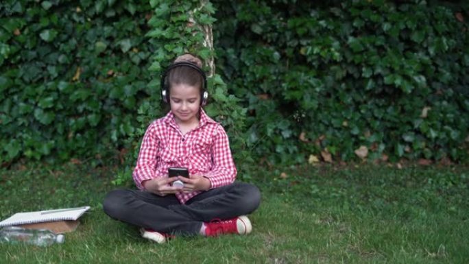 在阳光明媚的日子里，一名大学生坐在草坪上使用智能手机的肖像。高加索高中生女孩在大学公园里戴着耳机听音