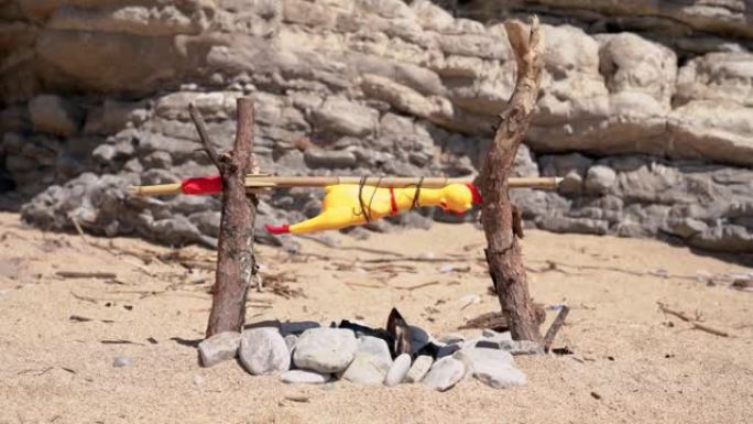 野生原住民准备篝火烤绑在自制唾沫上的玩具鸡。在荒岛上迷路的人。狩猎的概念。野餐，海滩上温暖的晴天