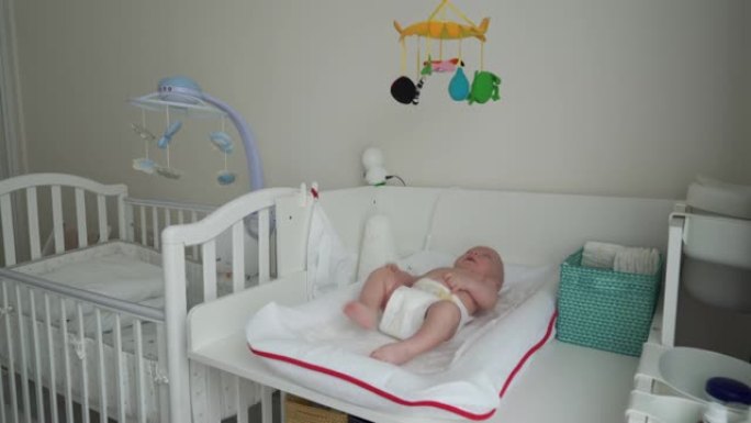 穿着尿布的婴儿在白色换衣桌上看着他上方的玩具手机，即3个月大的男婴