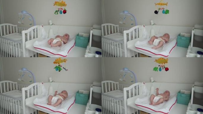 穿着尿布的婴儿在白色换衣桌上看着他上方的玩具手机，即3个月大的男婴