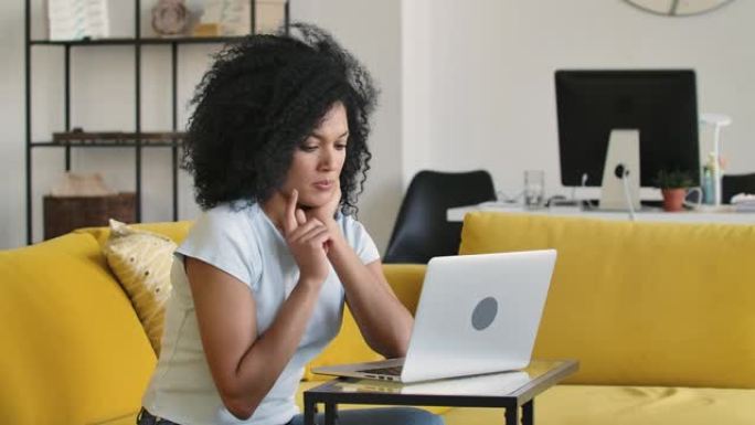 一个年轻的非洲裔美国妇女在笔记本电脑键盘上打字的肖像。黑发，卷发，坐在明亮的家庭房间里的黄色沙发上。