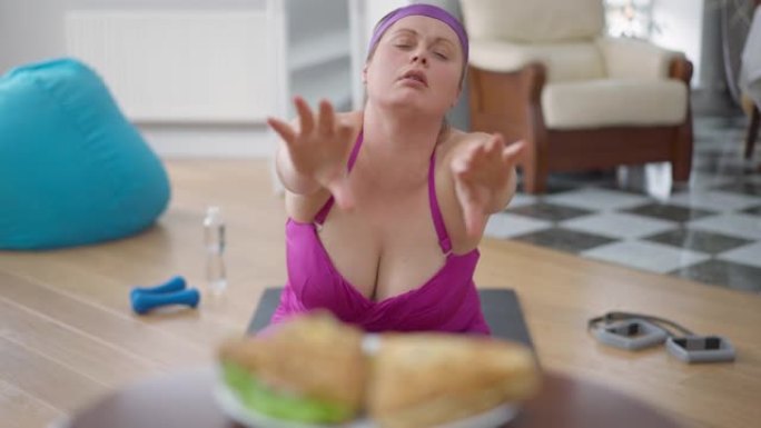 年轻的肥胖妇女抽水压榨机伸手去拿三明治。超重的白人女士在家室内训练。运动与灵感