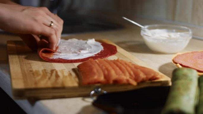 薄薄的美味彩色煎饼。女人的手的特写镜头，将鲑鱼片放在木板上涂上奶油奶酪的红色煎饼上。滚动煎饼。4k视
