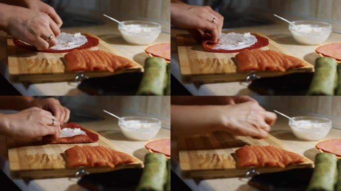 薄薄的美味彩色煎饼。女人的手的特写镜头，将鲑鱼片放在木板上涂上奶油奶酪的红色煎饼上。滚动煎饼。4k视