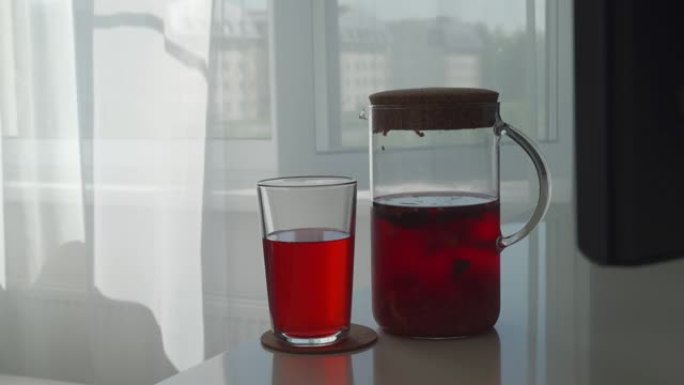 自制清爽的冷红色水果饮料，在醒酒器和玻璃中，在阳光下有新鲜的草莓薄荷柠檬水，窗户上有透明的白色窗帘