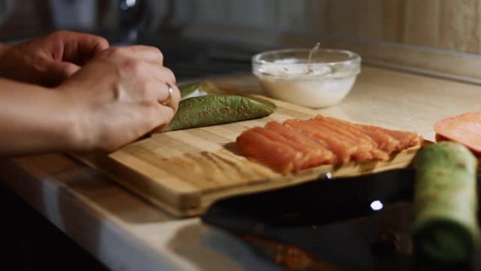薄薄的美味彩色煎饼。女人的手放在鲑鱼片上的特写镜头，填充在木板上涂上奶油奶酪的绿色煎饼上。滚动煎饼。